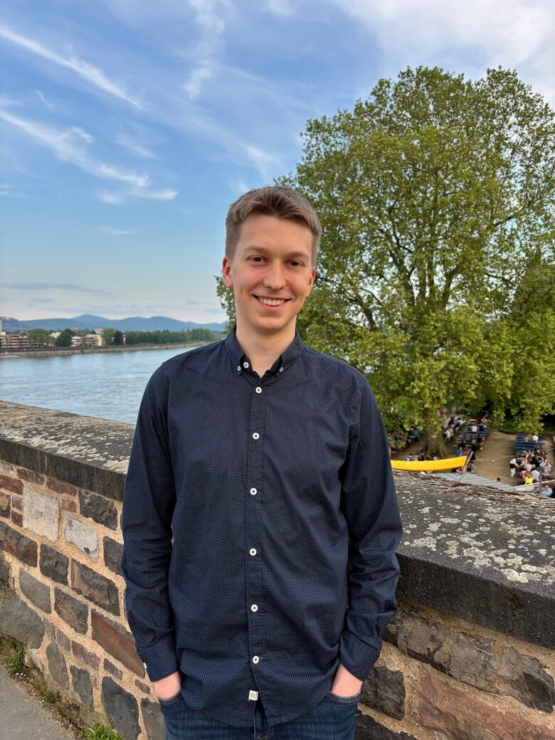 Lucas Brenner stands in front of a small wall with the Rhine and the mountains of the Siebengebirge in the background